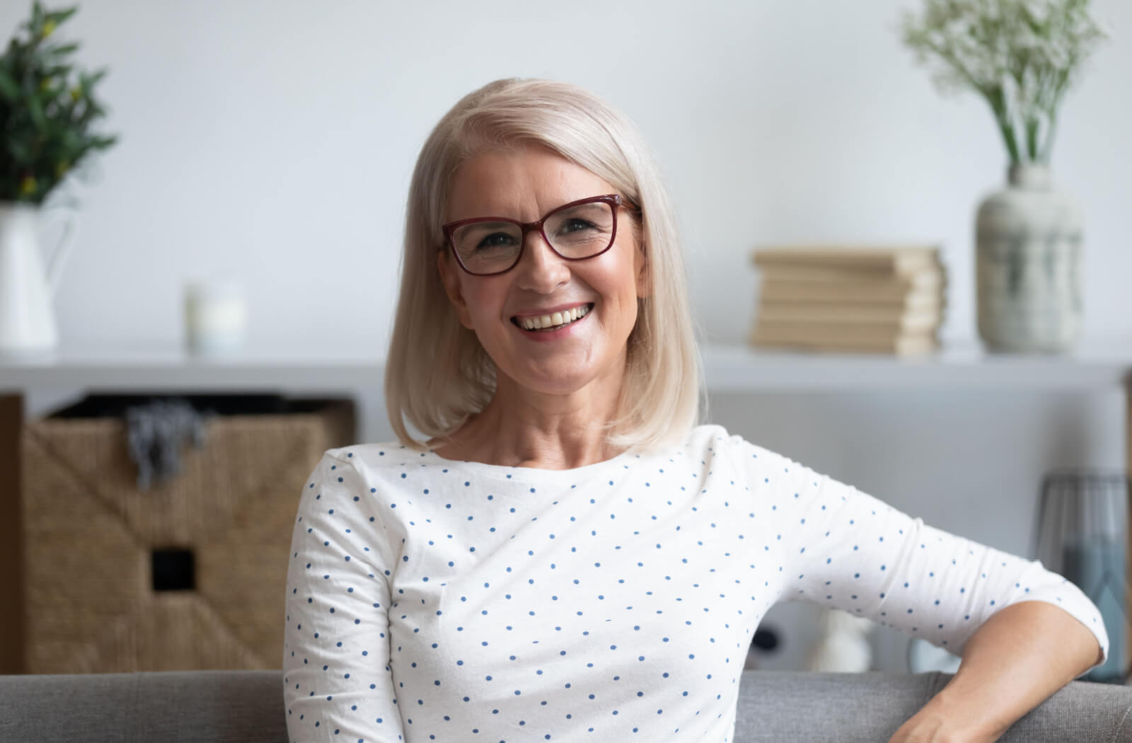 A senior woman with short white hair is smiling and sitting in a couch as she looks directly at the camera