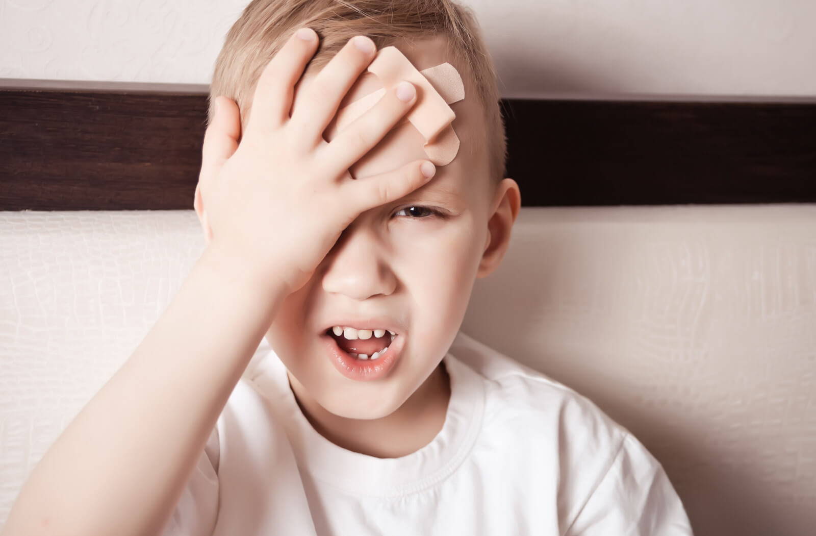 Close-up image of a young boy with 2 band-aids on his forehead. He is holding his right hand over his right eye and making a pained expression